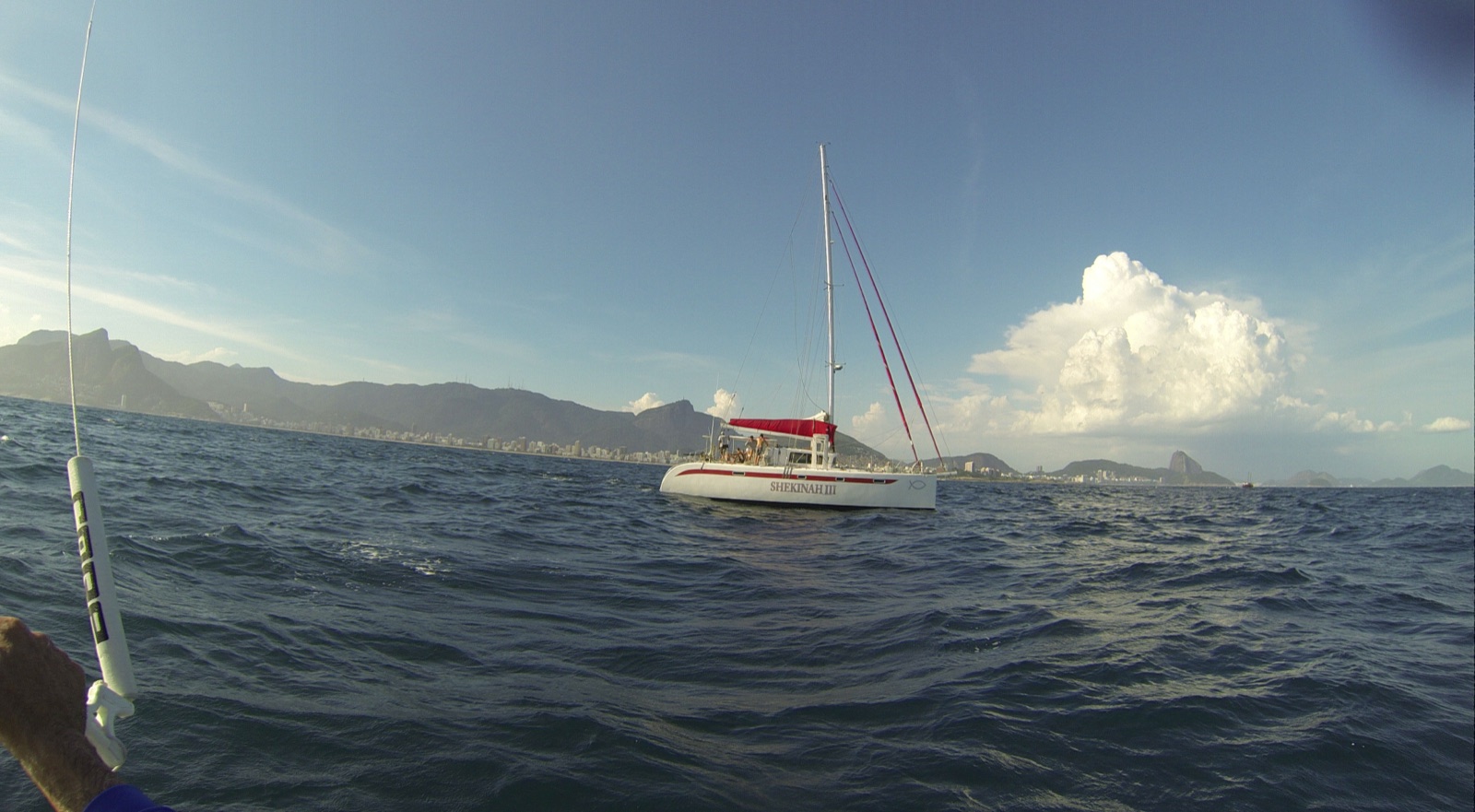 Catamarã de escolta em navegação ao largo de Ipanema, com a Pedra da Gávea e o Pão de Açúcar ao fundo